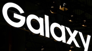 A glowing Samsung Galaxy logo stands out against a dark background. The image prominently features the large white letters "Galaxy," which run diagonally across the picture. In the background, only a few dark structures and small points of light are visible.