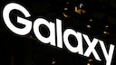 A glowing Samsung Galaxy logo stands out against a dark background. The image prominently features the large white letters "Galaxy," which run diagonally across the picture. In the background, only a few dark structures and small points of light are visible.