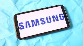 A smartphone lies horizontally on a light blue, crumpled surface. The display shows the blue Samsung logo in large capital letters against a white background. The device features a dark frame with a slightly bronze-colored edge. The smartphone is positioned at an angle in the image and occupies most of the frame.