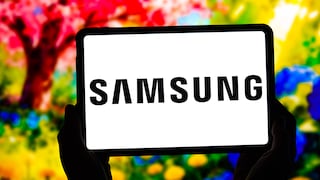 A tablet is held with both hands against a blurred, colorful background. The black Samsung logo is prominently displayed in the center of the white screen. In the background, vibrant shades of red, yellow, green, and blue are visible.