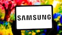 A tablet is held with both hands against a blurred, colorful background. The black Samsung logo is prominently displayed in the center of the white screen. In the background, vibrant shades of red, yellow, green, and blue are visible.
