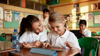 Two elementary school children sit together at a table in a classroom, looking at a tablet. In the background, more children and a teacher can be seen. A water bottle is also on the table.