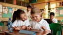 Two elementary school children sit together at a table in a classroom, looking at a tablet. In the background, more children and a teacher can be seen. A water bottle is also on the table.
