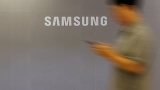 A large Samsung logo is displayed on a light gray wall. To the right, a blurred figure is seen holding a smartphone and moving past the wall. The image is intentionally designed with motion blur, emphasizing the white Samsung logo as the focal point.