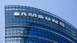 Close-up of the upper floors of a modern office building with a curved glass facade. Large white letters spelling "SAMSUNG" are affixed to the facade. The sky in the background is clear and blue.