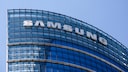 Close-up of the upper floors of a modern office building with a curved glass facade. Large white letters spelling "SAMSUNG" are affixed to the facade. The sky in the background is clear and blue.