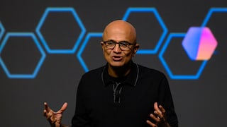 Satya Nadella, wearing glasses and a dark top, stands on a stage with both hands slightly raised. In the background, several blue hexagonal line graphics and a blurred hexagonal symbol in blue and magenta on a dark background are visible.