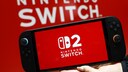 A person holds a Nintendo Switch 2 with the startup screen on against a red background featuring the Nintendo Switch logo. The console is shown from the front and is supported from below with one hand.