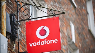 A red Vodafone sign with a white logo and lettering hangs on a black metal bracket attached to a brick facade. Above the bracket, pigeon deterrent spikes are installed, and in the background, other parts of the building are visible but out of focus.