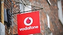 A red Vodafone sign with a white logo and lettering hangs on a black metal bracket attached to a brick facade. Above the bracket, pigeon deterrent spikes are installed, and in the background, other parts of the building are visible but out of focus.