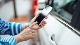 A person holds an iPhone in front of the driver's door of a white car and taps on the screen. The smartphone is positioned right next to the door handle, with more vehicles blurred in the background.