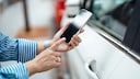 A person holds an iPhone in front of the driver's door of a white car and taps on the screen. The smartphone is positioned right next to the door handle, with more vehicles blurred in the background.