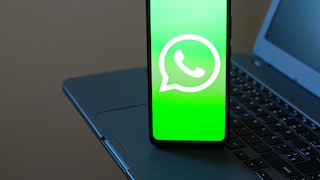 A smartphone rests on a laptop keyboard. The bright green display shows the white WhatsApp logo. In the background, parts of the laptop are visible but out of focus.