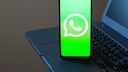 A smartphone rests on a laptop keyboard. The bright green display shows the white WhatsApp logo. In the background, parts of the laptop are visible but out of focus.
