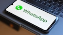 A smartphone with the WhatsApp startup screen open is positioned at an angle on a laptop keyboard. The display shows the green WhatsApp logo and the word "WhatsApp" on a white background. The black keys of the keyboard fill the background of the image.