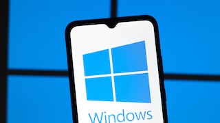Smartphone with a prominently displayed Windows logo on the screen set against a blurred blue background reminiscent of the typical Windows logo windows. The device is slightly tilted in the foreground, while the blue lighting in the background echoes the Windows theme.