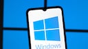 Smartphone with a prominently displayed Windows logo on the screen set against a blurred blue background reminiscent of the typical Windows logo windows. The device is slightly tilted in the foreground, while the blue lighting in the background echoes the Windows theme.