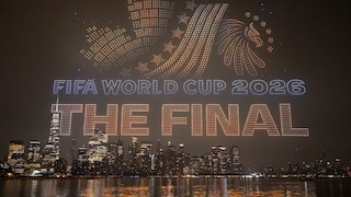 Drone Show Over a Nighttime Skyline by the Water: Hundreds of illuminated drones form the words "FIFA WORLD CUP 2026" and "THE FINAL," along with a soccer motif in the sky. In the background, numerous lit-up skyscrapers and their reflections on the water's surface are visible.