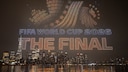 Drone Show Over a Nighttime Skyline by the Water: Hundreds of illuminated drones form the words "FIFA WORLD CUP 2026" and "THE FINAL," along with a soccer motif in the sky. In the background, numerous lit-up skyscrapers and their reflections on the water's surface are visible.