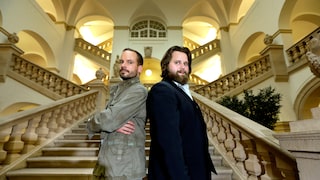 Alt-text: Two people stand back-to-back on a wide staircase in a brightly lit building with arches, railings, and tall windows.