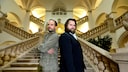 Alt-text: Two people stand back-to-back on a wide staircase in a brightly lit building with arches, railings, and tall windows.