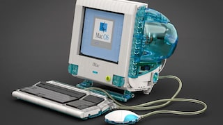 A Lego replica of an Apple iMac G3 sits against a dark background. The computer features a transparent blue case with an integrated screen displaying the Mac OS logo. In front of the computer are a matching keyboard and the round "hockey puck" mouse, both connected to the model with transparent cables.