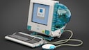 A Lego replica of an Apple iMac G3 sits against a dark background. The computer features a transparent blue case with an integrated screen displaying the Mac OS logo. In front of the computer are a matching keyboard and the round "hockey puck" mouse, both connected to the model with transparent cables.