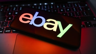 A smartphone is positioned at an angle on a laptop keyboard. The display shows the colorful eBay logo against a dark background. The keyboard and the laptop's casing are illuminated by red and violet light reflections. The background is blurred.