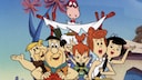 Animated scene from "The Flintstones": Several characters are sitting together in an open Stone Age vehicle. Two characters are visible in the front, one at the wheel and the other in the passenger seat, with more characters and a small child behind them. A pink dinosaur with a long neck towers over the vehicle. The car has a roof made of wooden poles and white fabric. In the background, stylized trees with violet-blue trunks and pink leaves stand against a light blue sky.