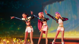 Three animated figures stand on a lit stage, performing a choreography. All three are dressed in short white outfits with black, glittering jackets and gold-colored details. The figure on the left extends an arm forward, the middle figure with long pink hair raises both arms, and the figure on the right with long violet hair holds a hand to her head. In the blurred foreground, lights and an audience are visible. The background features a colorfully lit concert setting in shades of blue, green, and yellow.