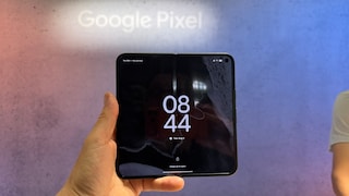 A hand holds an open Google Pixel 11 Pro Fold in front of a large backdrop with the words "Google Pixel." The inner display of the foldable smartphone shows the lock screen with the time 8:44. The crease in the middle of the display is visible. The background features shades of pink and blue, and on the right, a partial view of a person in a light-colored top can be seen.