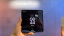 A hand holds an open Google Pixel 11 Pro Fold in front of a large background wall with the words "Google Pixel." The inner display of the foldable smartphone shows the lock screen with the time 8:44. The crease in the middle of the display is visible. The background features shades of pink and blue, and on the right, a partial view of a person in a light-colored top can be seen.