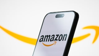 A smartphone is positioned at an angle against a bright background. The display shows the Amazon logo with black lettering and an orange arrow. In the background, a large, blurred orange Amazon arrow stretches across the image. The smartphone has a black frame and a pill-shaped cutout at the top of the display.
