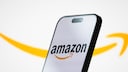 A smartphone is positioned at an angle against a bright background. The display shows the Amazon logo with black lettering and an orange arrow. In the background, a large, blurred orange Amazon arrow stretches across the image. The smartphone has a black frame and a pill-shaped cutout at the top of the display.