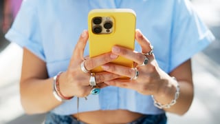 A person is holding a yellow smartphone with three camera lenses on the back in both hands. The person is wearing a light blue top, several rings on their fingers, and bracelets on their wrists. The background is blurred and brightly lit.