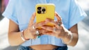 A person is holding a yellow smartphone with three camera lenses on the back in both hands. The person is wearing a light blue top, several rings on their fingers, and bracelets on their wrists. The background is blurred and brightly lit.