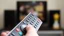 A hand holds a remote control pointed at a television. The TV in the background is blurred, displaying an unidentifiable program on the screen. Speakers are positioned to the left and right of the television.