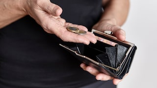 A person holds an open black wallet in one hand and several euro coins in the other. The wallet contains a few bills, and the coin compartment appears largely empty. The background is blurred and bright.