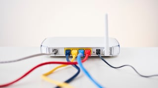 A white Wi-Fi router sits on a light surface against a neutral background. The back features several ports with network cables plugged in, colored blue, yellow, light blue, and red. To the right, there's a single white antenna, next to which a black cable is connected. The cables extend forward out of the frame and are partially blurred.