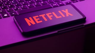 Alt-text: A smartphone with the red Netflix logo is placed diagonally on a laptop keyboard. The image is captured in violet-pink lighting.