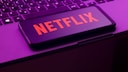 Alt-text: A smartphone with the red Netflix logo is placed diagonally on a laptop keyboard. The image is captured in violet-pink lighting.