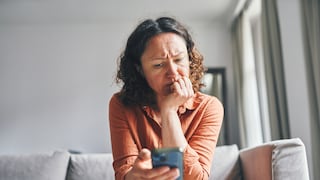 A person sits on a light-colored sofa in a living room, holding a smartphone in their right hand. They lean forward, resting their chin on their left hand. In the background, a window with curtains and a mirror are visible.
