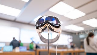 Apple Vision Pro on a metal stand in an Apple Store. The smart glasses are centered in the image, with several people standing between wooden tables in the background. The sales area is brightly lit, and the surroundings appear blurred. A cable runs from the glasses to a device on the table.