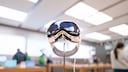 Apple Vision Pro on a metal stand in an Apple Store. The smart glasses are centered in the image, with several people standing between wooden tables in the background. The sales area is brightly lit, and the surroundings appear blurred. A cable runs from the glasses to a device on the table.