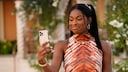 A person with long dark hair holds a smartphone in front of them outdoors. They are wearing a sleeveless top with a red and black pattern, large hoop earrings, and gold-colored jewelry. In the blurred background,