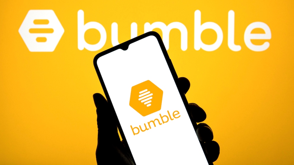 Against a yellow background, the white Bumble logo is prominently displayed. In the foreground, a hand holds a smartphone, which also shows the Bumble logo on its bright screen.