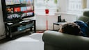 A person is lying on a sofa, looking at a TV in a bright living room. The screen displays various streaming content in a menu view. In front of the window, there's a small round table with a plant in a red pot. Additional furniture and electronic devices are situated next to the sofa and TV. Daylight streams through the large windows into the room.