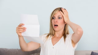 A person sits on a sofa holding a piece of paper. One hand rests on their head. A bright wall is visible in the background. The document is being examined closely.