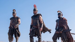 Three individuals in armor that appears ancient stand on a sandy hill under a clear blue sky. The person in the center wears a helmet with a red plume, a cloak, and a sword at their side. The two individuals on the left and right also wear helmets, breastplates, and greaves. In the background, several tents and more figures can be seen on a sandy encampment. The scene is bathed in bright daylight.