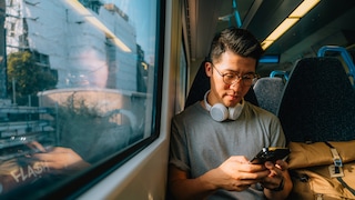 A person sits by the window on a train, holding a smartphone. White headphones hang around their neck. The window reflects the person as buildings and railway facilities pass by outside. A light-colored backpack rests on the adjacent seat.